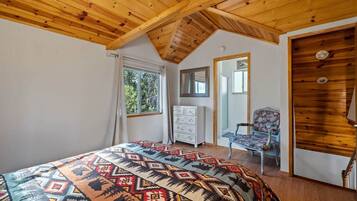 Une chambre avec un plafond en bois incliné, un lit recouvert d’une couverture à motifs, une commode, une chaise et un miroir accroché au mur.