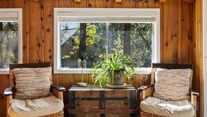 A cozy room with wooden walls and a large window. There are two wicker chairs with cushions, a vintage trunk, and a potted plant on a wooden table. - Walk to town, Lake access, Pet Friendly, King bed (Twain Harte)
