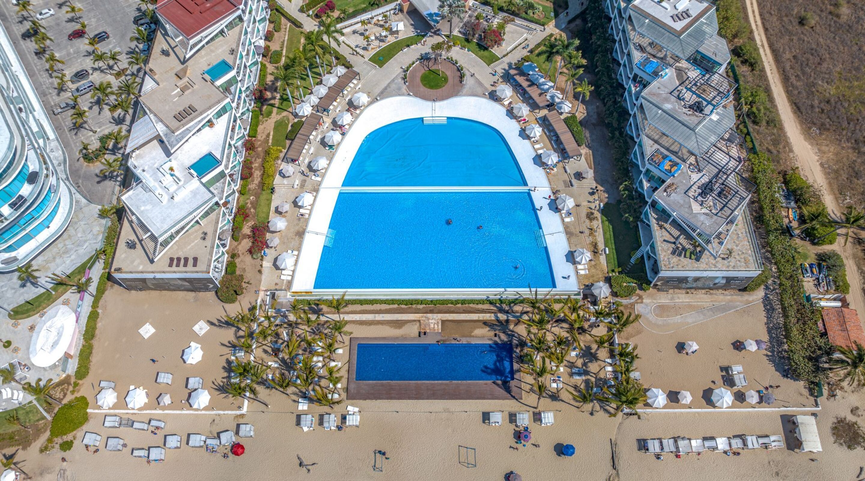 Ovenfra set et resort med en central swimmingpool, flere loungeområder ved stranden og en parkeringsplads.