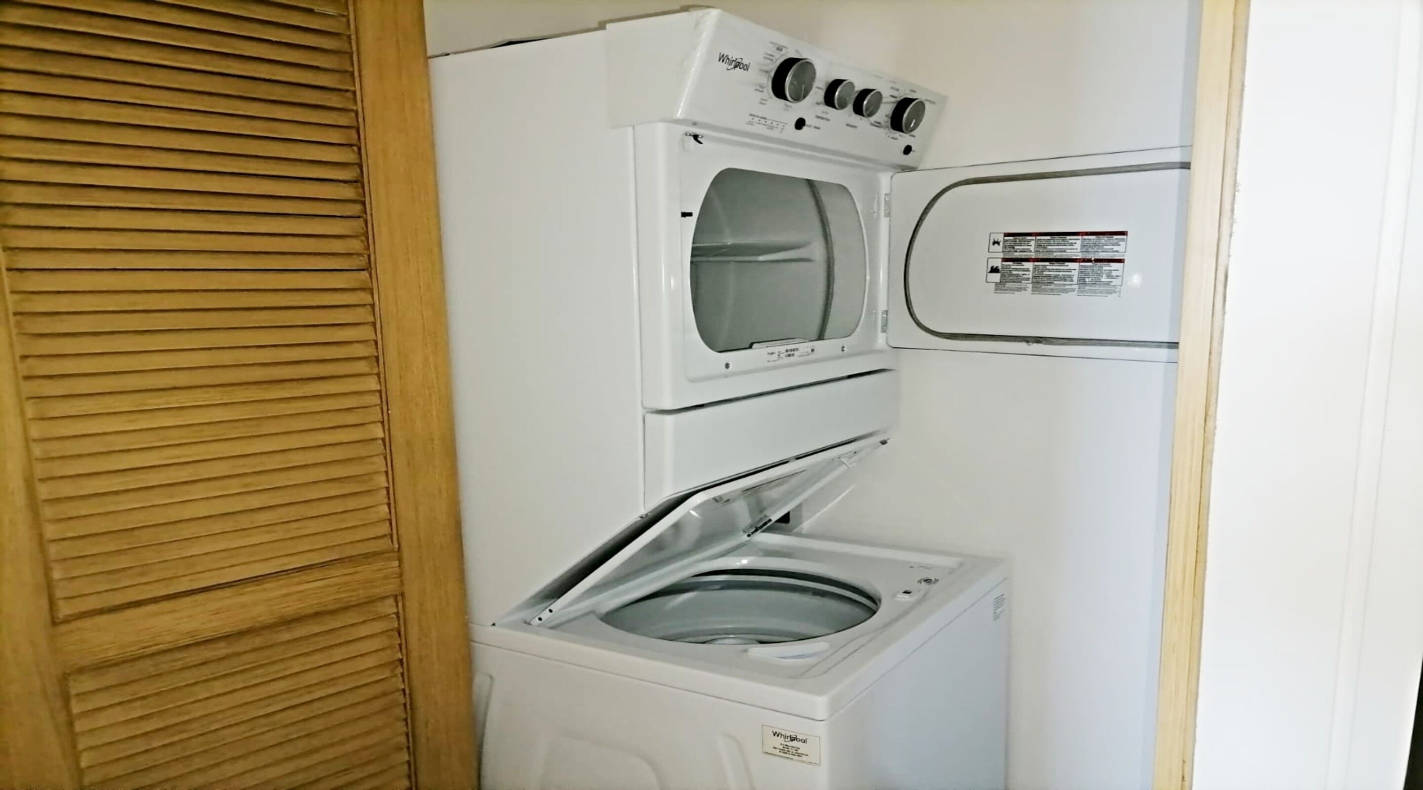 Een kleine wasruimte met een voorlader wasmachine en droger, houten jaloezieën en een klein raam.