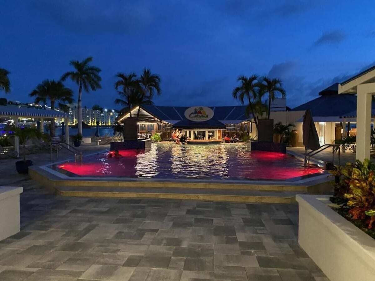 A swimming pool area with a water feature, palm trees, and a building with a logo in the background.
