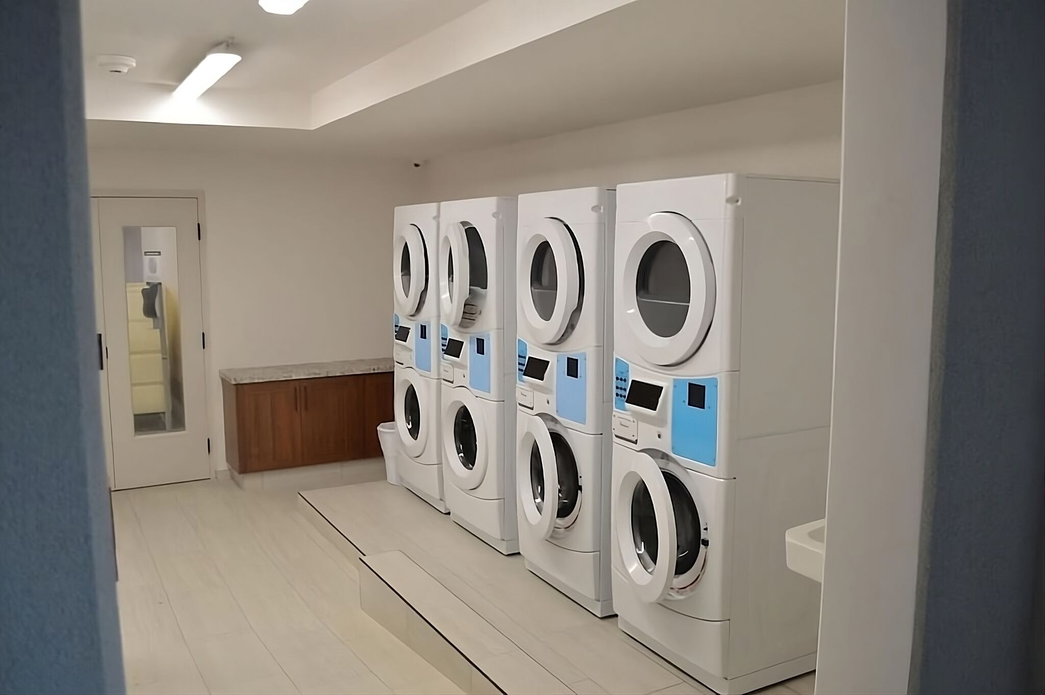 A laundry facility with multiple washing machines, a wooden bench, and a white door.
