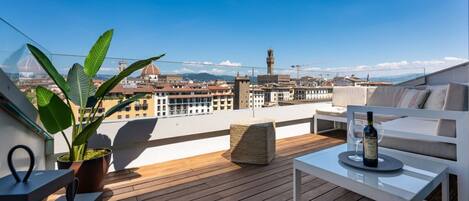 A rooftop terrace with a view of a cityscape, including a dome and tower, furnished with a sofa, table, and potted plant.
