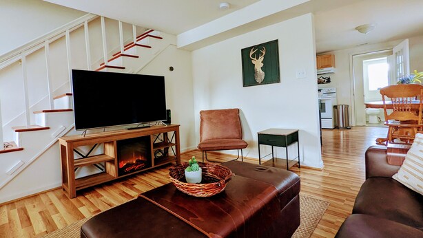 Ein Wohnzimmer mit Fernseher auf einem Holzschrank, einer braunen Ledersofa, einem kleinen Tisch mit Korb und einer Treppe mit roten Geländern.