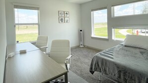Ett hotellrum med en säng, ett skrivbord, två stolar, en portabel värmare och stora fönster med utsikt över omgivningarna.