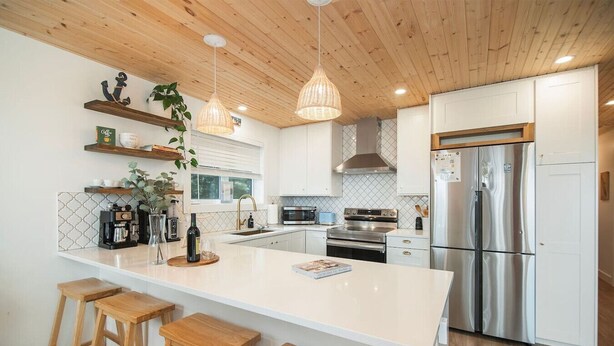 Une cuisine moderne dotée d’un îlot central, de tabourets en bois, d’appareils électroménagers en acier inoxydable et de luminaires suspendus.
