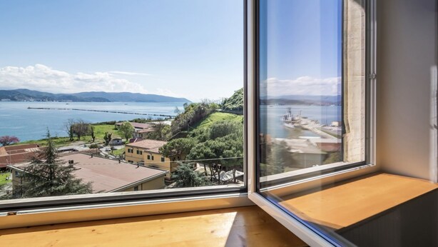 A room with a large window offering a view of a coastal area, including a body of water, hills, and a distant cityscape.
