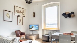 A modern living room with a large window, a flat-screen TV, a dining area, and artwork on the walls.