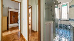A hallway leading to a bedroom with a wooden door, a bathroom with a shower and tiled floor, and a view of a bedroom with a bed and a window.