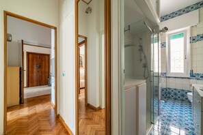 A hallway leading to a bedroom with a wooden door, a bathroom with a shower and tiled floor, and a view of a bedroom with a bed and a window. - GuestHost - Cozy apartment of 90 sqm, can accommodate up to 5 people, located on the second floor of a building with a lift (NOT suitable for disabled people - 6 steps to access). The accommodation is located near one of the venues of the Sapienza Univer (Roma)