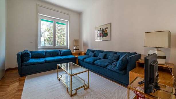 A living room with two blue sofas, a glass coffee table, a TV, and a window with blinds.