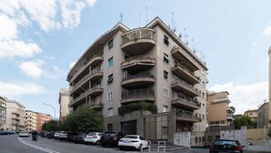 A multi-story residential building with balconies, a gated entrance, and parked cars along the street.