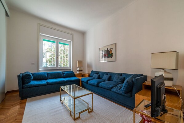Una sala de estar con dos sofás azules, una mesa de centro de vidrio, un televisor y una ventana con persianas.