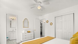 Un dormitorio con cama, ventilador de techo, armario, espejo y baño visible a través de una puerta abierta.