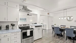 Una cocina moderna con armarios blancos, estufa de acero inoxidable y un comedor con espejo redondo y luces colgantes.