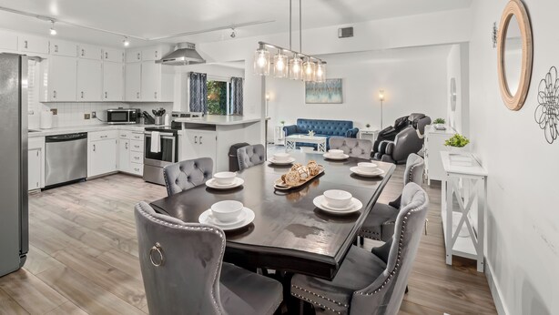 Una cocina moderna con armarios blancos, un comedor con mesa de madera oscura y sillas tapizadas en gris, y un sofá azul al fondo.