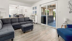 Una sala moderna con un sofá seccional, un reposapiés y un sillón azul. Hay un comedor con mesa y sillas, y una puerta corrediza de vidrio que da a un patio exterior.
