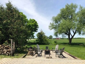 Une terrasse avec des fauteuils Adirondack, une table et un foyer, le tout entouré d’arbres et d’un champ d’herbe.
