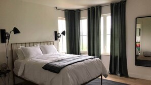 A bedroom with a bed, two bedside lamps, a window with curtains, and a mirror on the wall. - Sister Bay Eclectic Cottage (Sister Bay)
