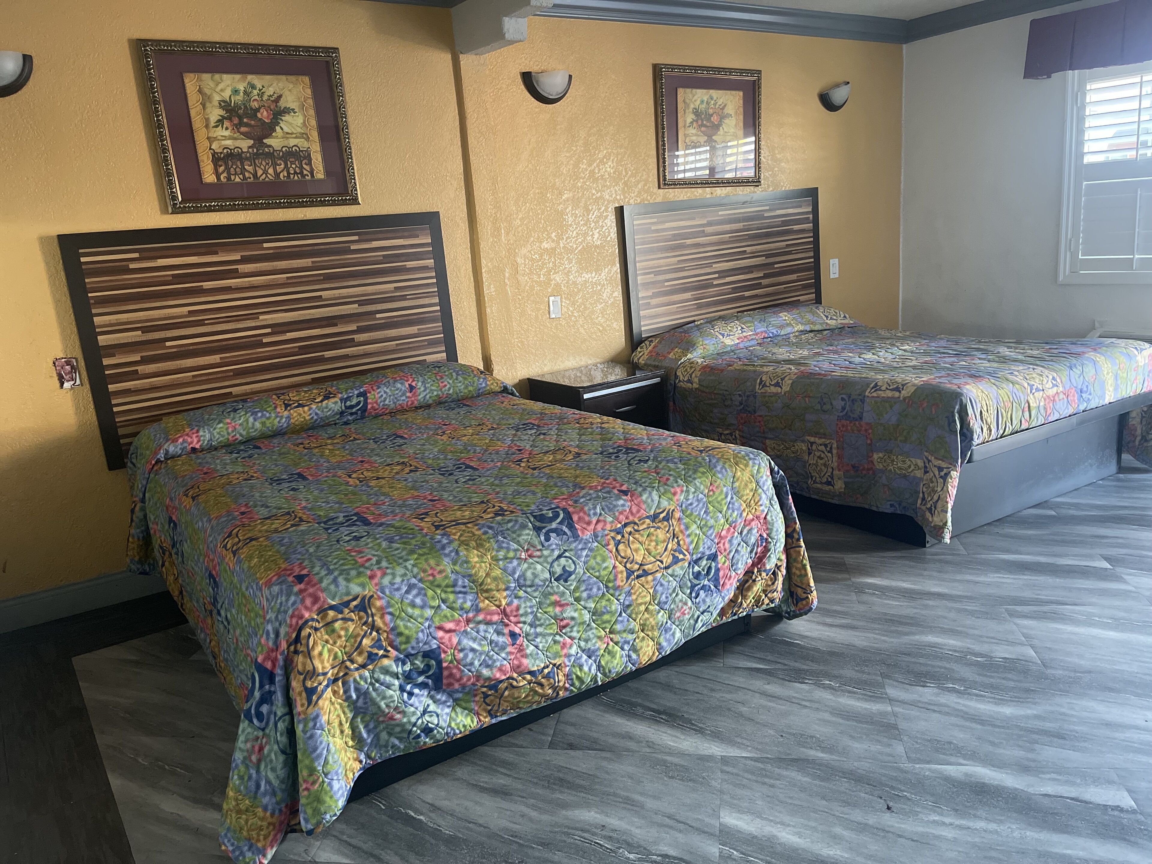 A hotel room with two beds, wooden headboards, floral-patterned bedding, and wall-mounted artwork.