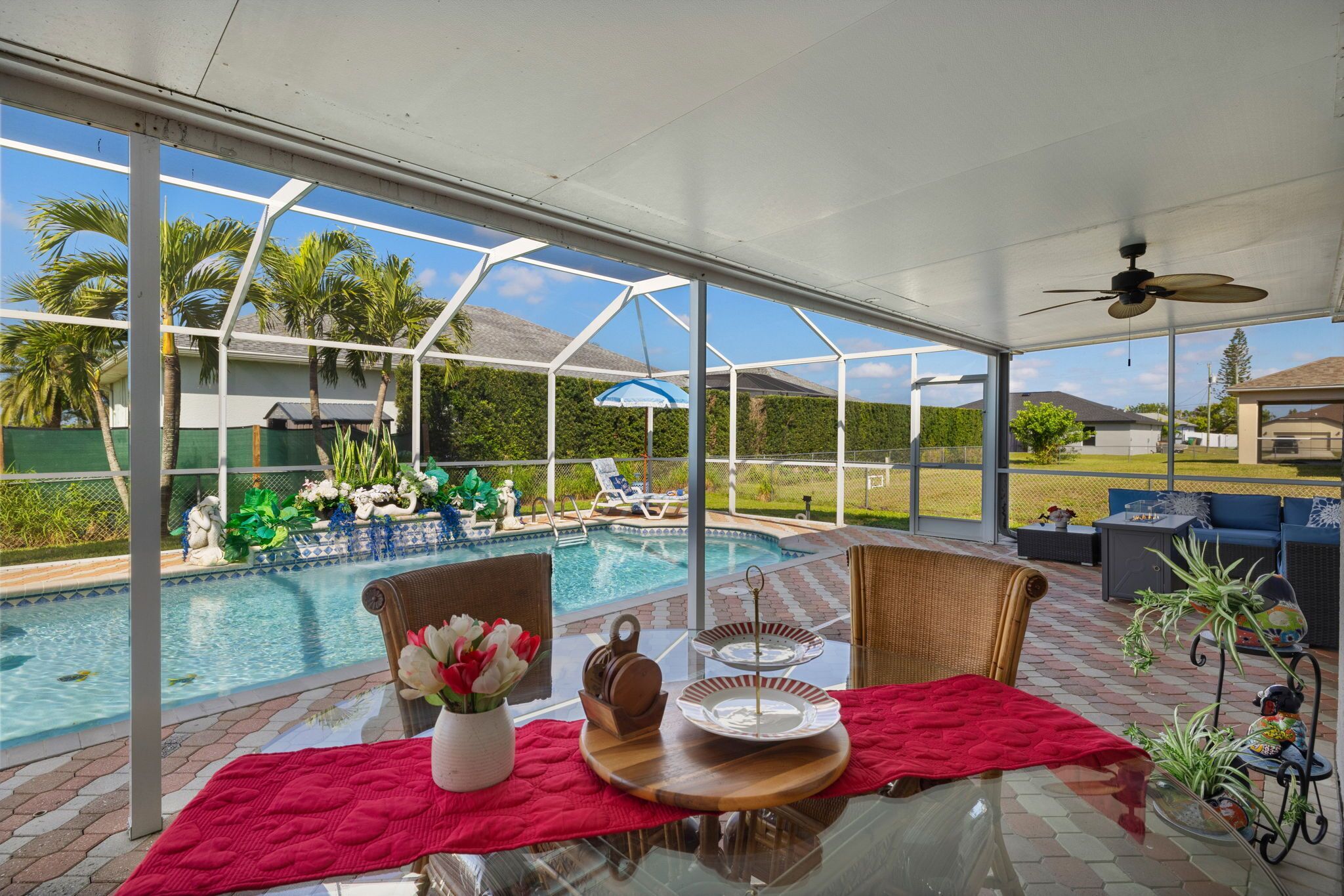 A covered outdoor area with a pool, lounge chairs, and a table set with a red table runner, a vase of flowers, and a tray.