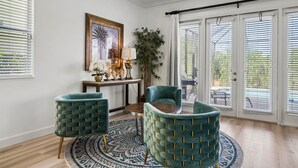 Une pièce avec une table ronde, deux fauteuils turquoise, une table d’appoint en bois avec une lampe, un tableau encadré et une grande plante en pot.