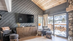 Una sala de estar moderna con techo de madera, un televisor de pantalla plana, un sofá, una mesa de centro y vistas a un paisaje nevado a través de amplios ventanales.