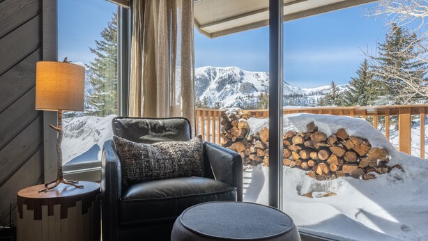 Una habitación acogedora con un sofá de cuero, un taburete redondo y una lámpara. Un ventanal grande ofrece vistas de montañas nevadas y una terraza de madera con leña apilada.