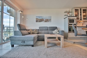 Ein modernes Wohnzimmer mit einem grauen Ecksofa, einem Holztisch und einem großen Fenster mit Blick ins Freie.