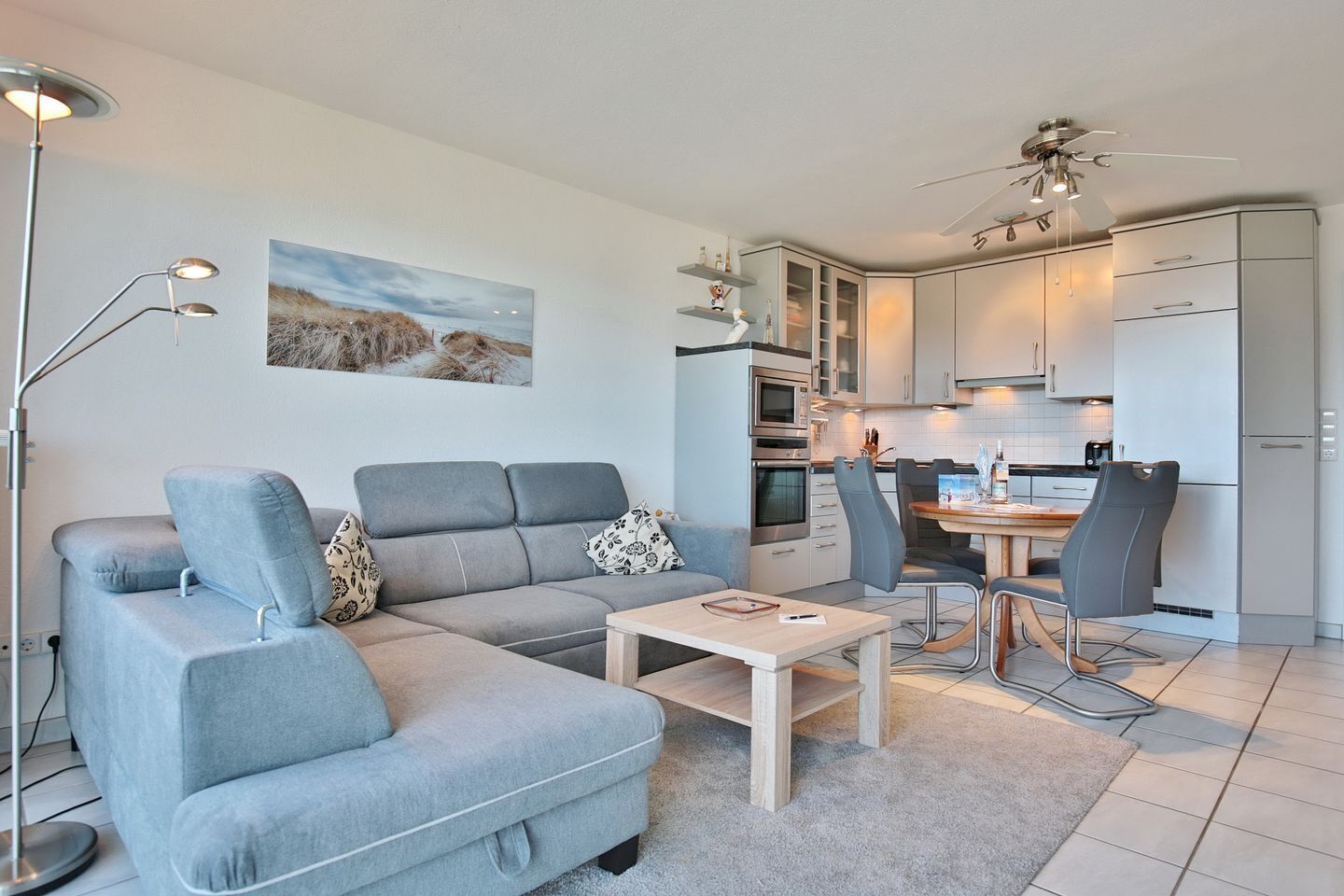 Ein modernes Wohnzimmer mit grauem Sofa, Couchtisch und Essbereich inklusive Tisch und Stühlen. Es gibt eine Küche mit weißen Schränken und einem Deckenventilator.