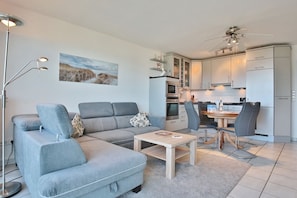 Ein modernes Wohnzimmer mit grauem Sofa, Couchtisch und Essbereich inklusive Tisch und Stühlen. Es gibt eine Küche mit weißen Schränken und einem Deckenventilator.