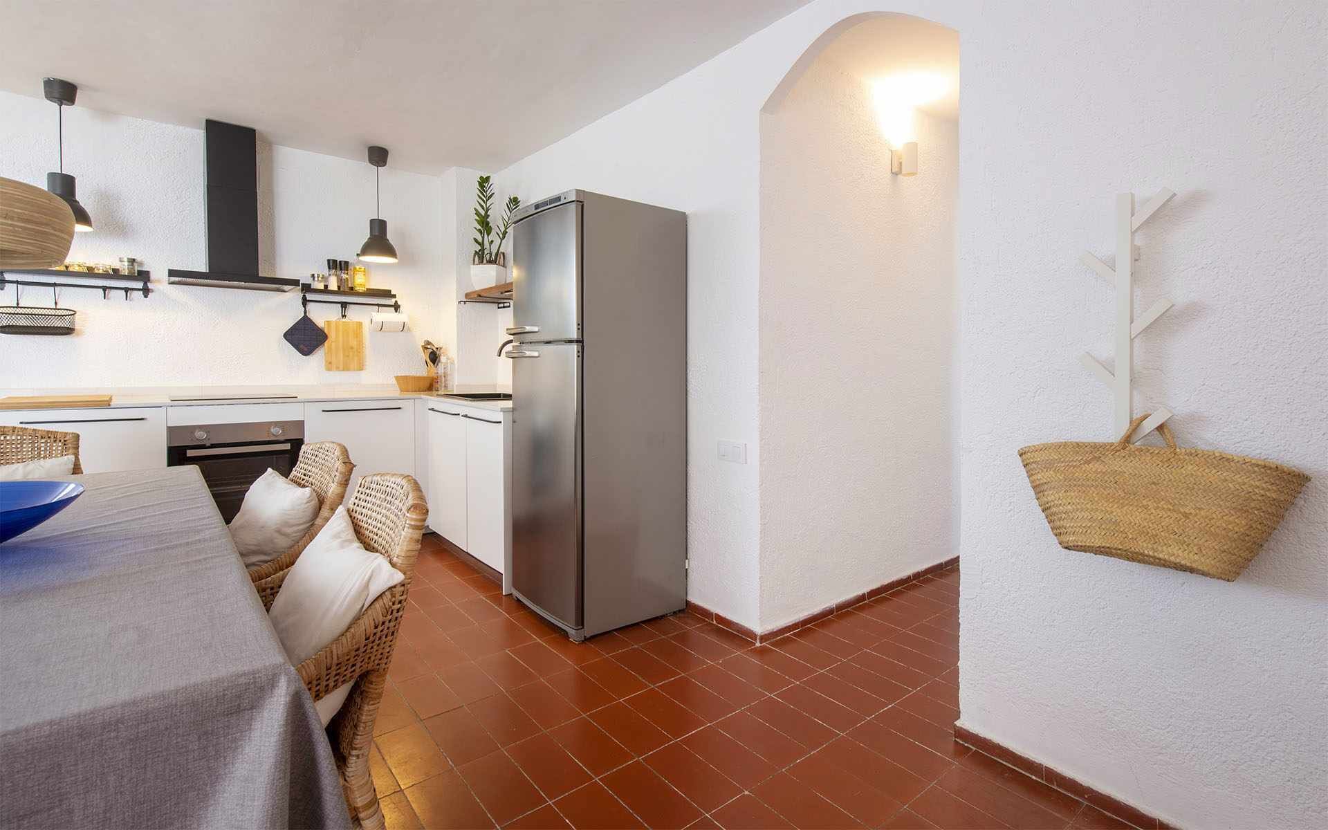 A compact kitchen with white cabinets, a stainless steel refrigerator, and a dining area with wicker furniture.