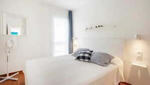 Une chambre avec un lit, un ventilateur sur pied, une fenêtre avec des rideaux, une étagère avec de petites plantes et un luminaire fixé au mur.