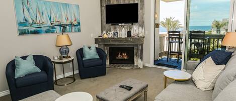 A living room with a fireplace, a television, and a balcony with a view of the ocean.