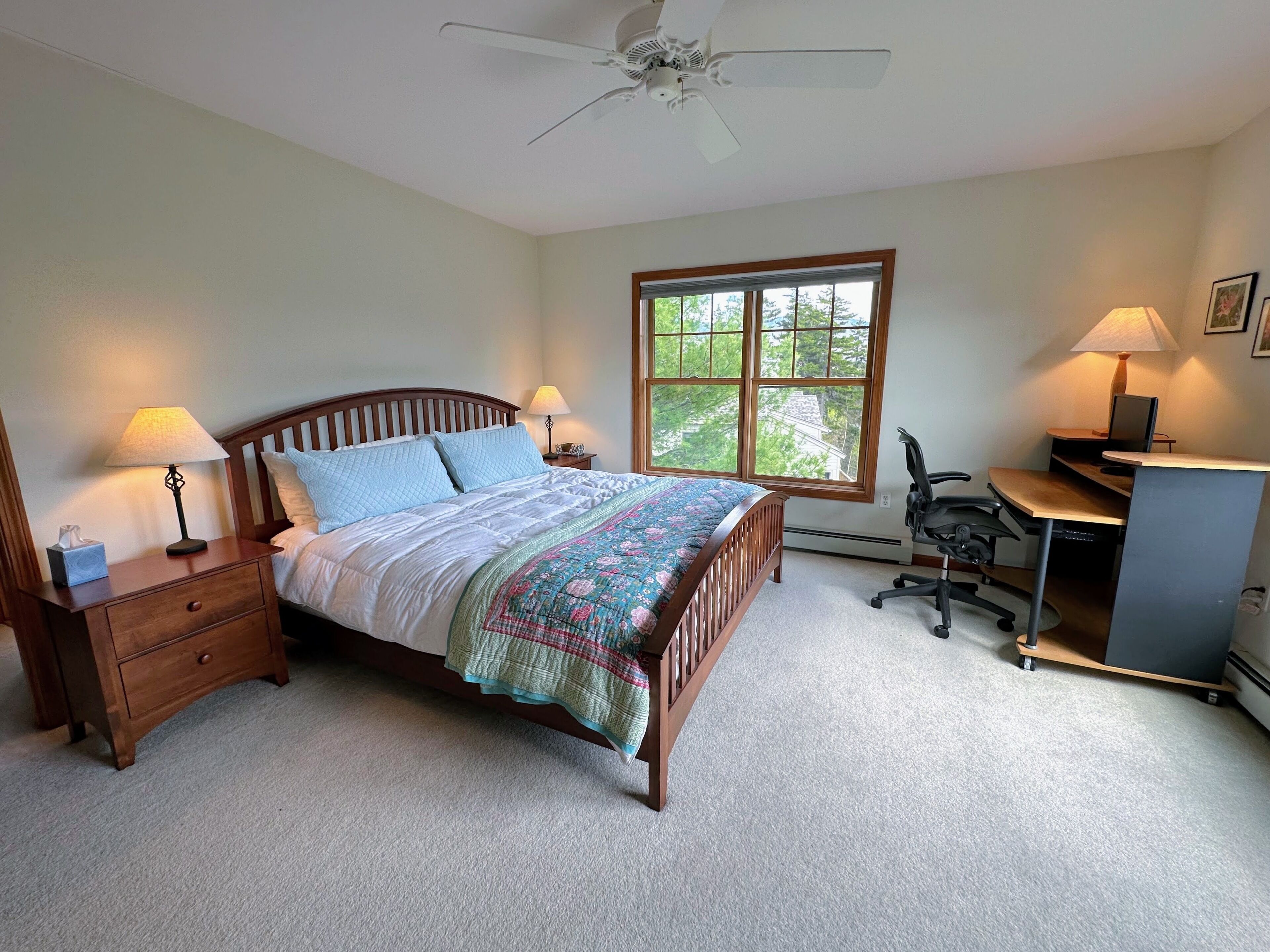 A bedroom with a bed, bedside lamps, a desk, a chair, and a window with a view of greenery.