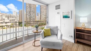 Una sala de estar moderna con un ventanal grande, un sofá gris con un cojín verde, una mesita redonda con una planta y vistas a la ciudad.