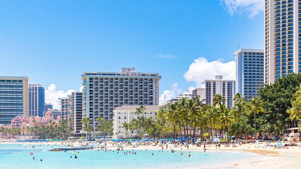 Un hotel frente a la playa con varios edificios altos, palmeras y una playa concurrida.