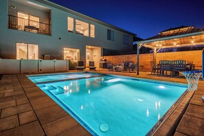 A swimming pool with a covered patio area, a barbecue setup, and a two-story building with windows.