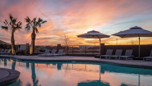 Zona de piscina con sillones de descanso y sombrillas, palmeras y un cielo al atardecer.