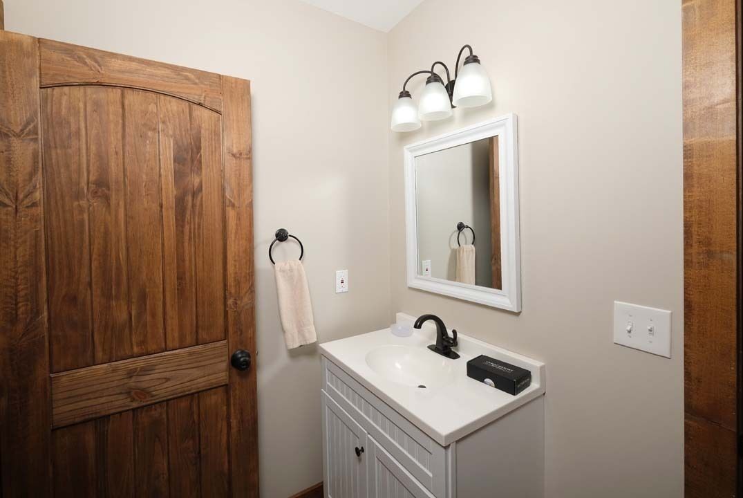 A bathroom with a wooden door, a white sink with a black faucet, and a mirror with three lights.