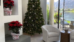 Un soggiorno moderno con un albero di Natale decorato, due poinsettie rosse in vaso e vista su un lago e montagne attraverso ampie finestre.