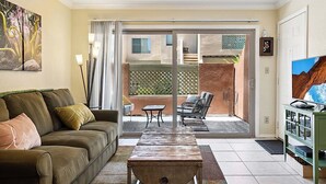 Una sala de estar con un sofá, una mesa de centro, un televisor y un ventanal corredera que da a un patio exterior con una silla y una mesa.