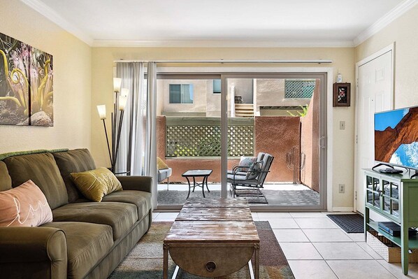 Un salon comprenant un canapé, une table basse, un téléviseur et une porte-fenêtre coulissante donnant sur une terrasse extérieure avec une chaise et une table.