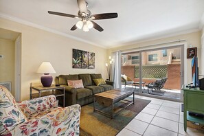 Un salon comprenant un canapé, une table basse et un ventilateur de plafond. Une porte-fenêtre coulissante donne accès à un espace extérieur aménagé avec une table et des chaises.