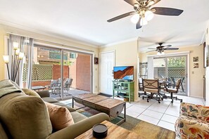 A spacious living room with a sofa, coffee table, and dining area with chairs. There is a sliding glass door leading to a patio with outdoor furniture.