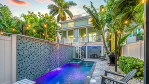 A swimming pool with a tiled wall, outdoor seating, and a two-story building with a balcony.
