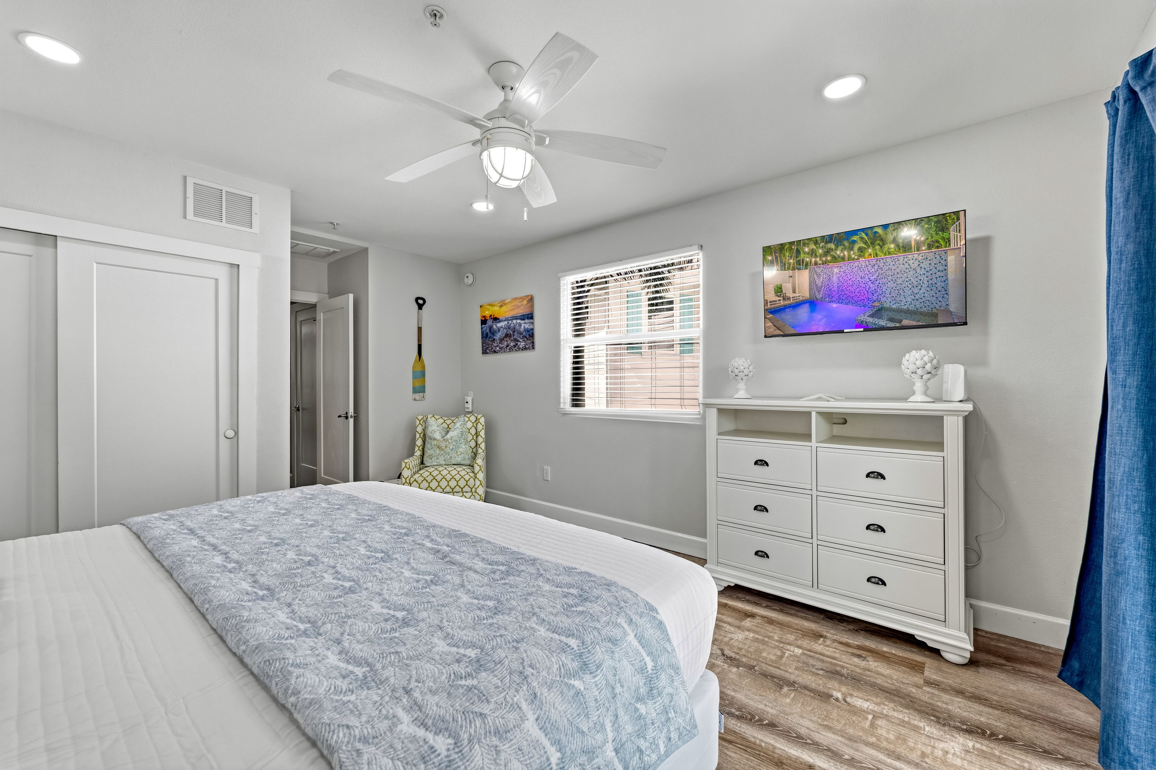 A bedroom with a bed, a ceiling fan, a window with blinds, a painting, and a white dresser.