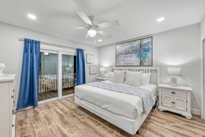 Une chambre à coucher comprenant un lit, des tables de chevet, un ventilateur de plafond, une grande fenêtre avec des rideaux et un tableau accroché au mur.