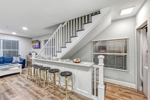 Un salon moderne avec un escalier, un canapé, une table basse et un coin cuisine équipé de tabourets.