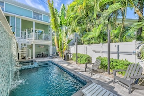 A swimming pool with a waterfall feature, surrounded by a patio with chairs and a barbecue grill.